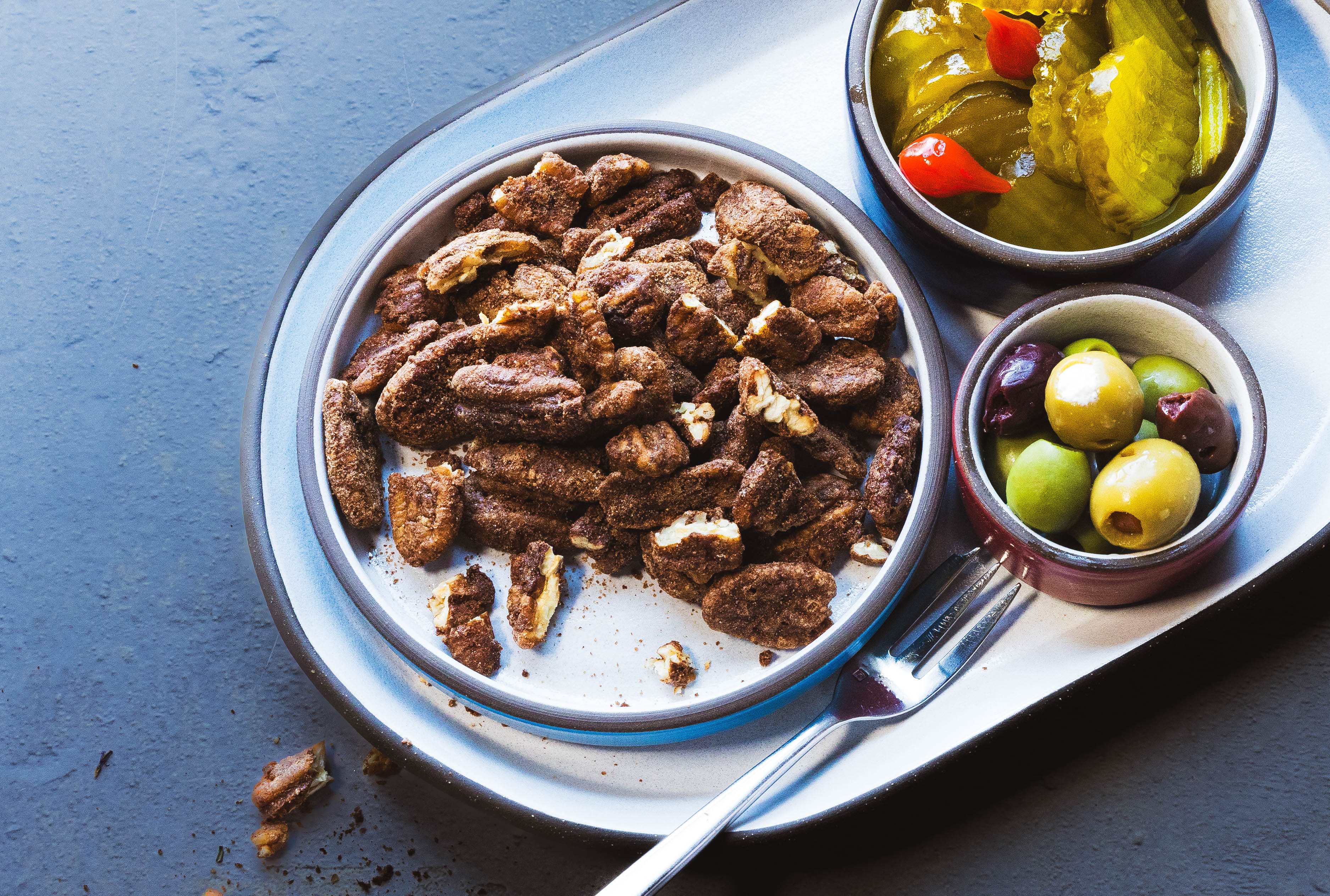 Tray with pecans seasoned with the House Blend, pickles, and olives on a blue surface