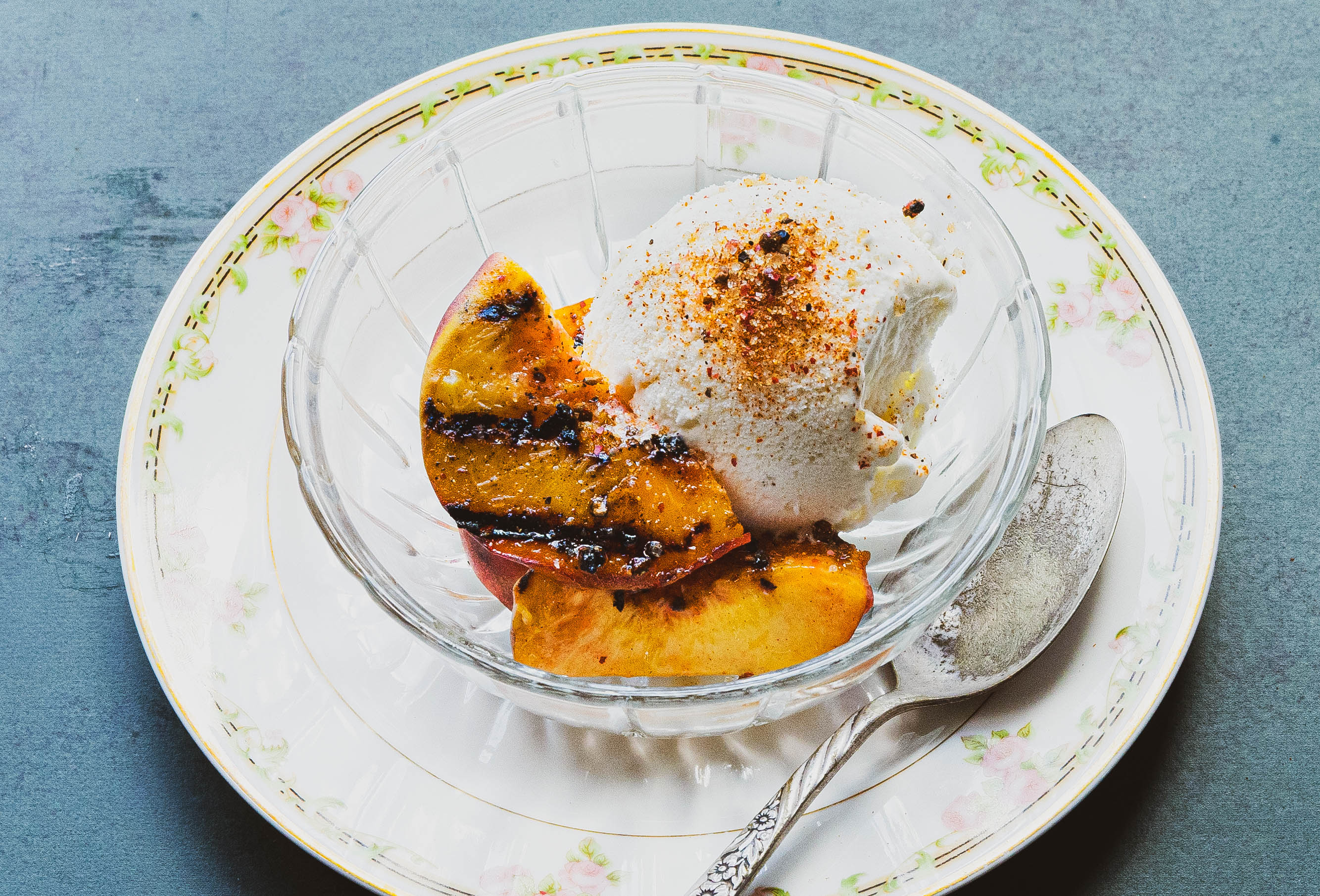 Glass bowl with grilled peaches using the Ember spice blend, and vanilla ice cream on a decorative plate with a spoon.
